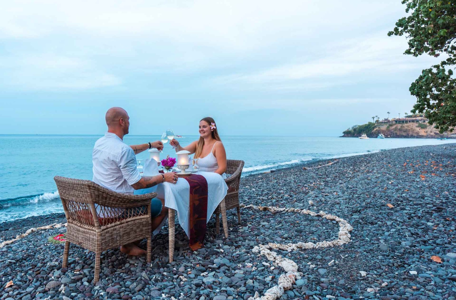 Couple toasting at a private candlelit dinner set on the black pebble beach inside a coral heart at Amed Lodge by Sudamala Resorts in Amed, east Bali