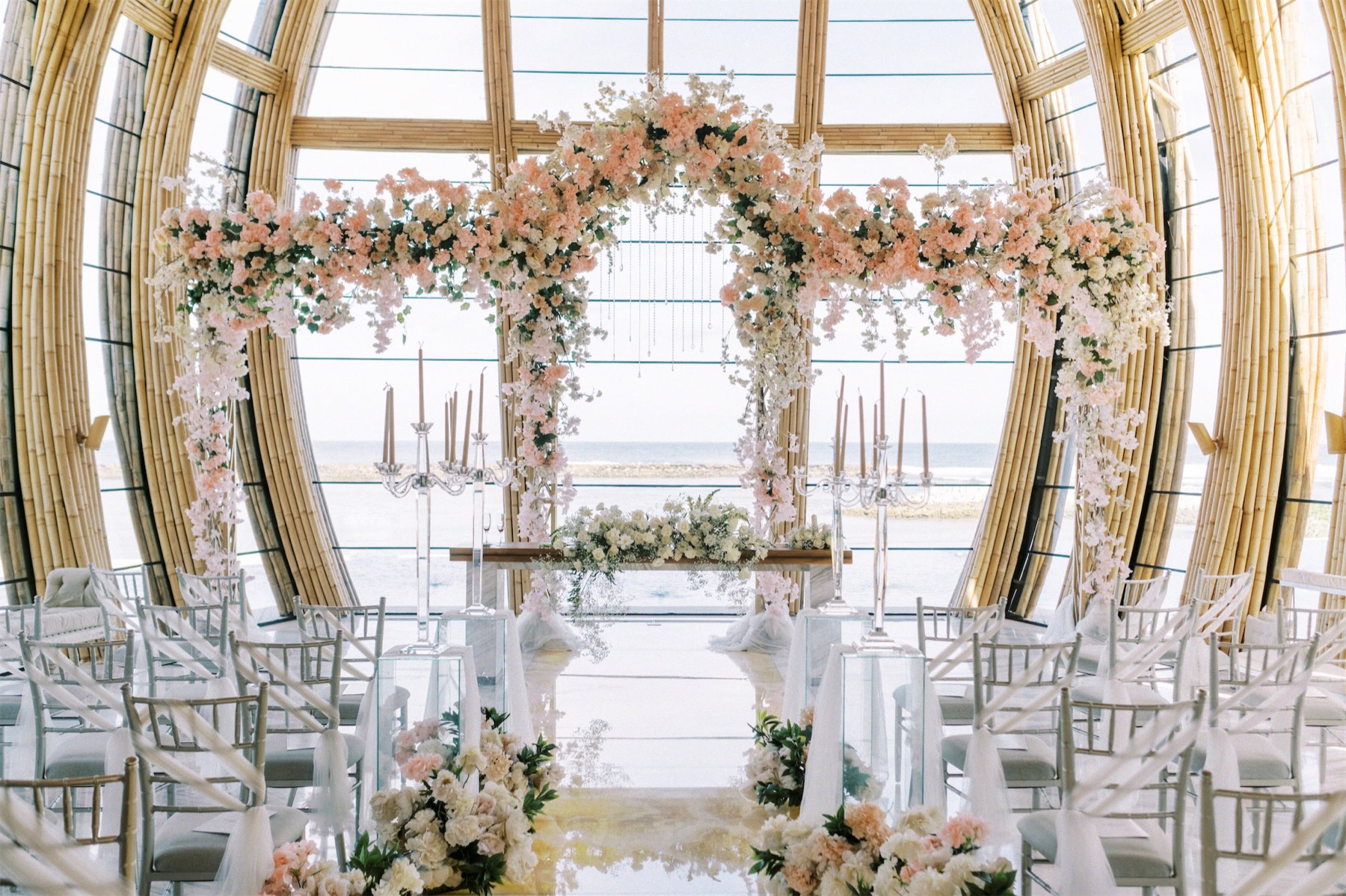 Ceremony inside the bamboo Apurva Chapel with a blush floral arch and ocean view, Nusa Dua