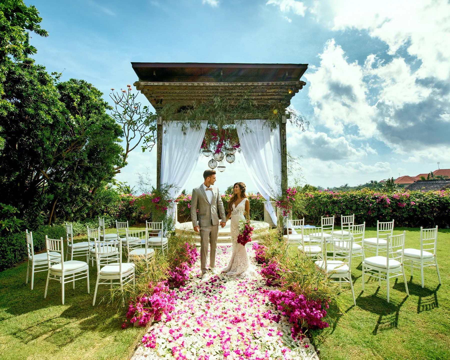 A couple beneath the carved timber joglo gazebo at Asmara Gazebo, AYANA, with white drapes, a bougainvillea aisle, and garden all around