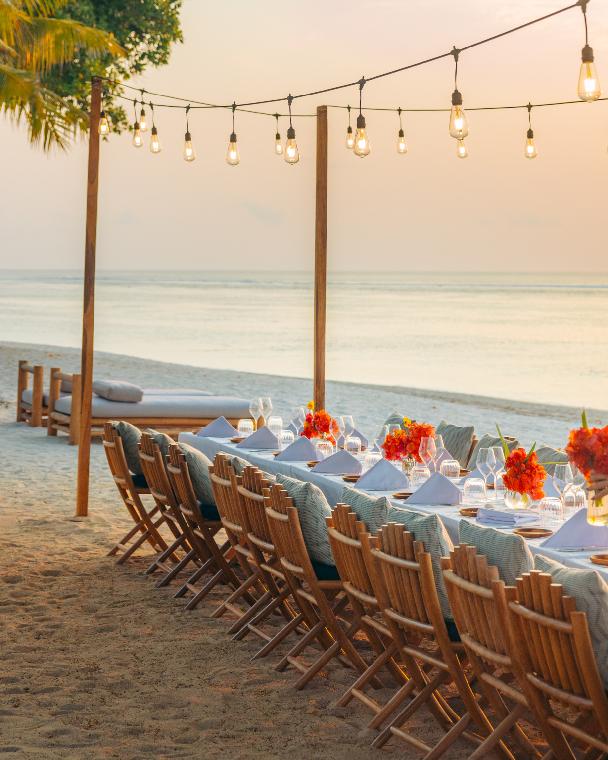 Beachfront banquet table with orange florals and hanging bulbs at dusk at Cap Karoso, Sumba