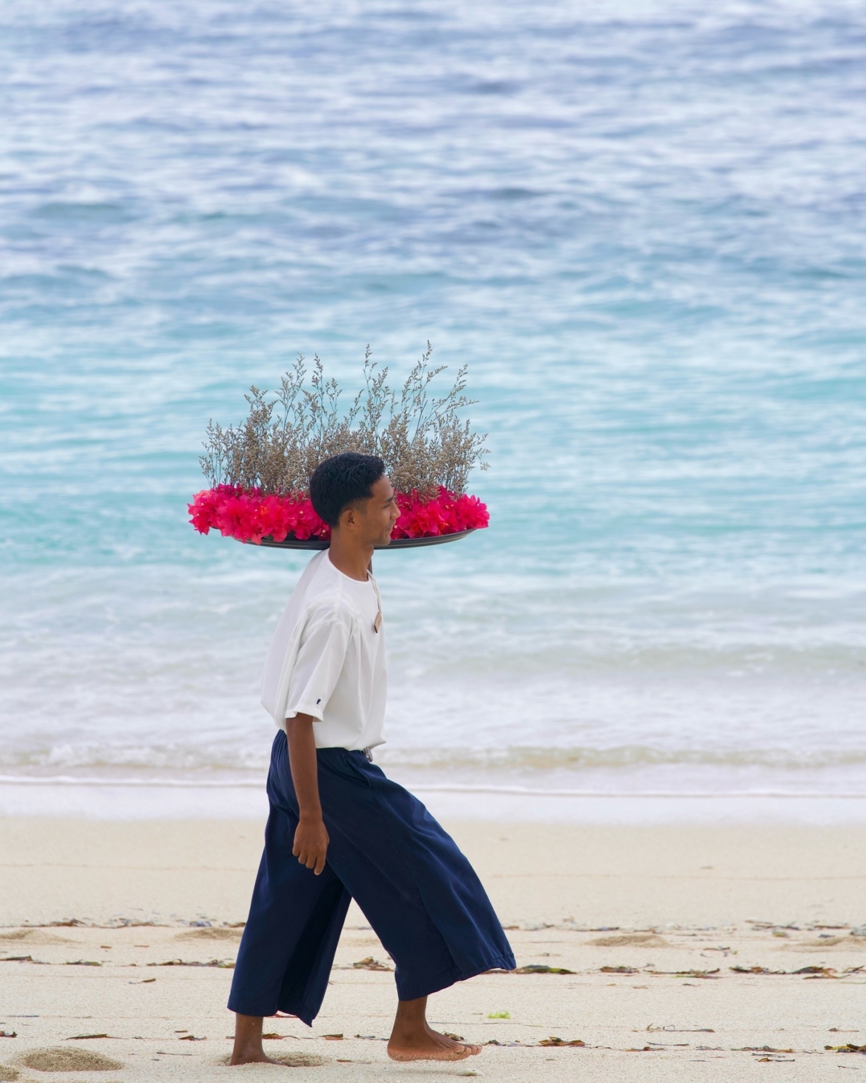 A waiter carrying a tray of pink bougainvillea on his head along the shoreline at Cap Karoso, Sumba