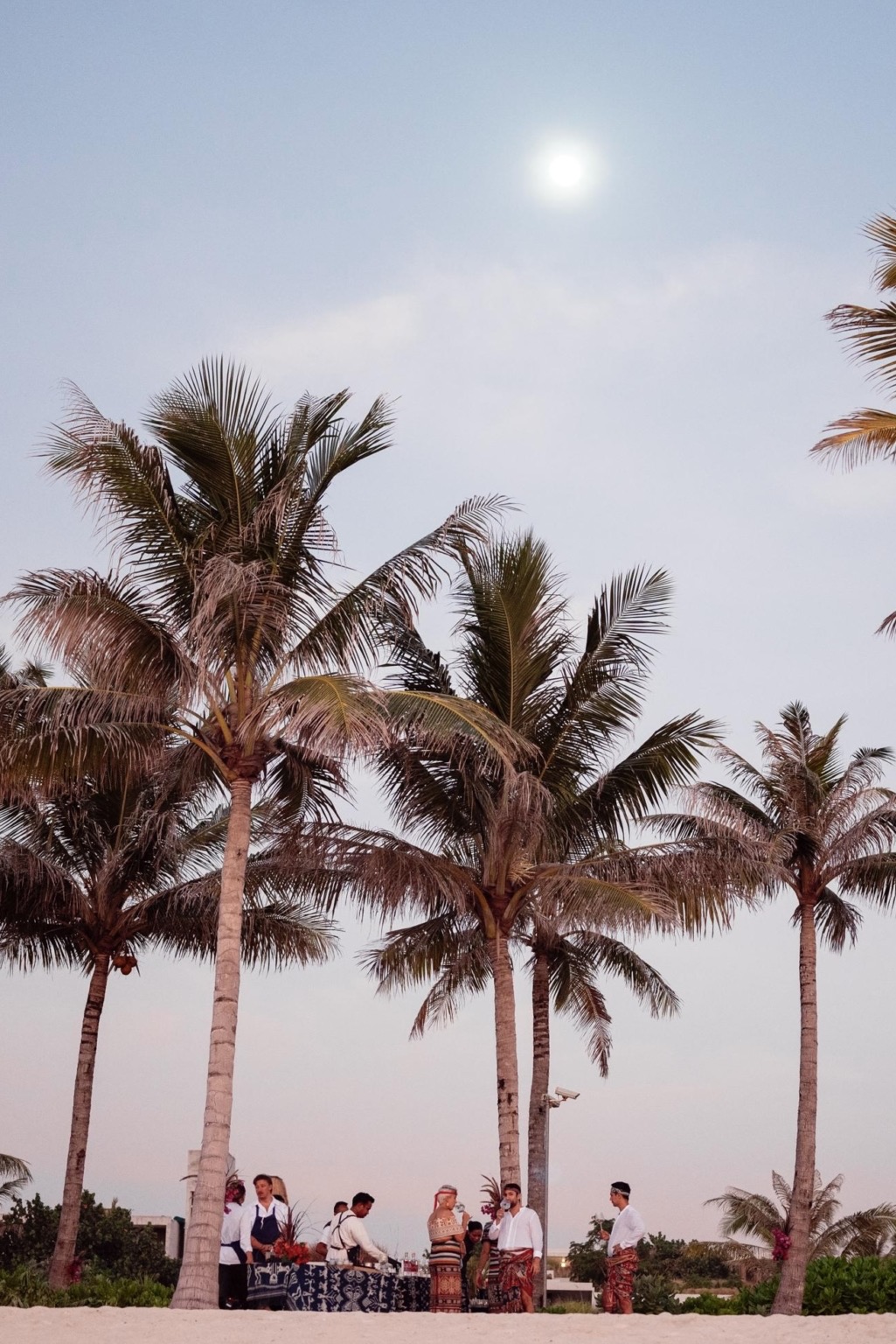 Staff in patterned sarongs setting up a beach bar beneath tall coconut palms at dusk at Cap Karoso, Sumba