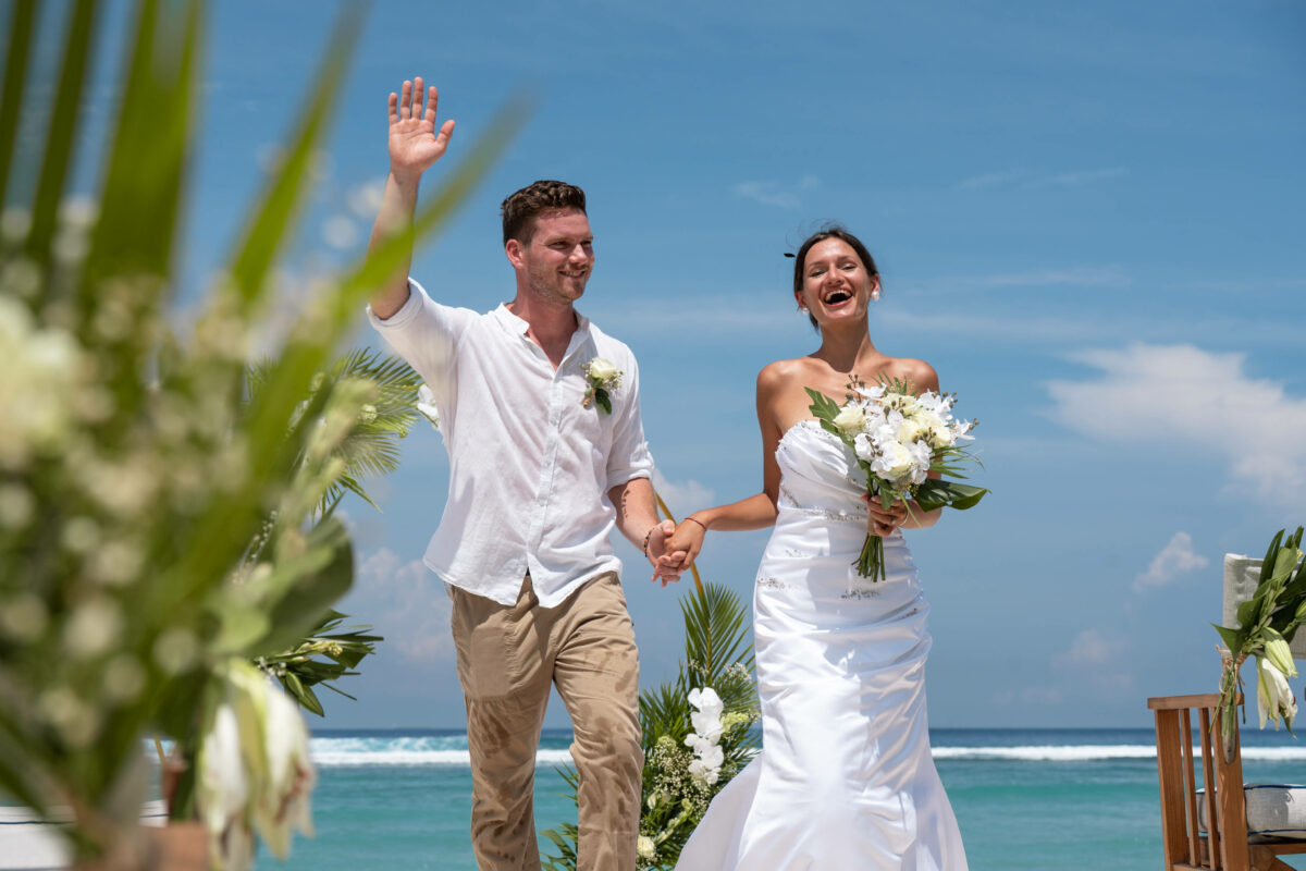 Newlyweds waving after their beach ceremony with the turquoise reef behind them at Cocana Resort, Gili Trawangan
