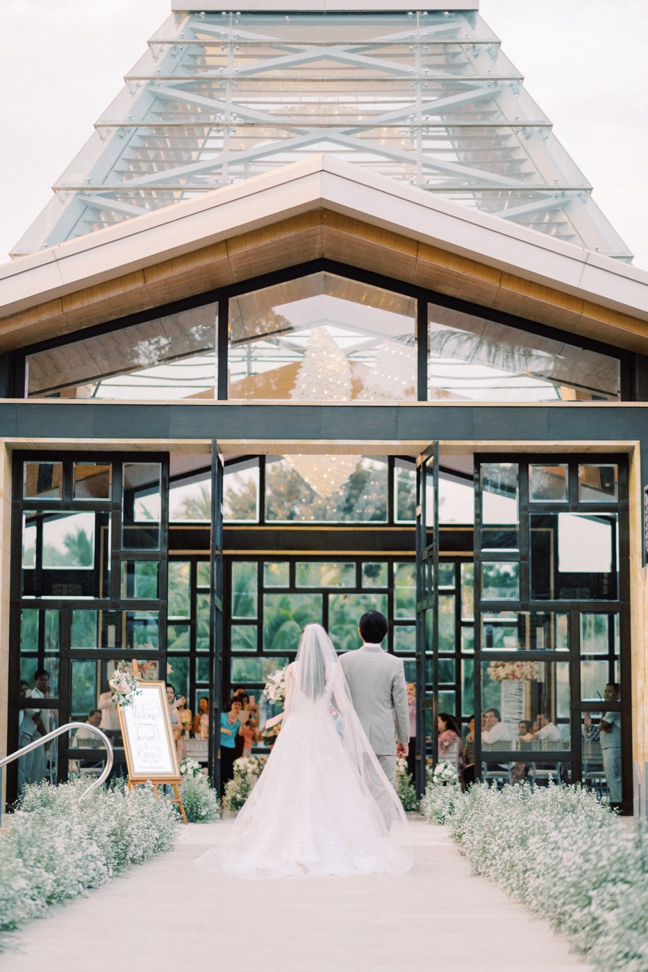 A couple entering the glass Eternity Chapel with its lantern-lit chandelier at The Mulia, Nusa Dua