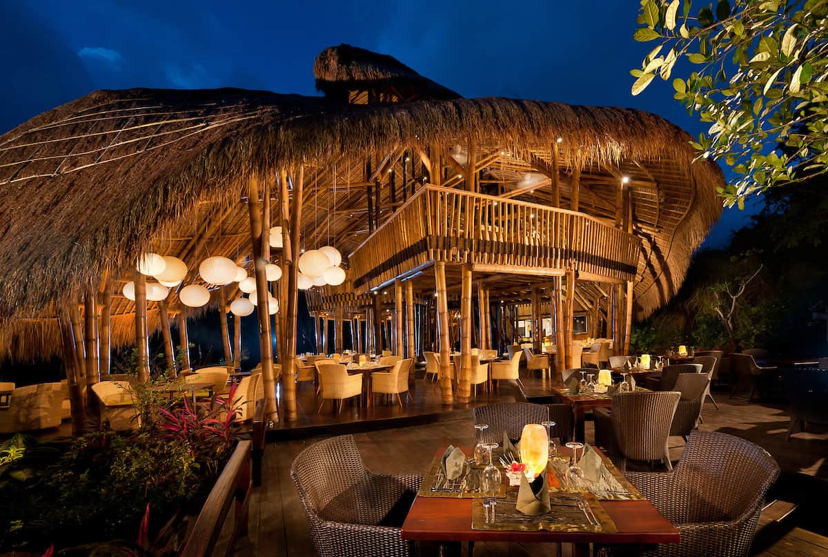 The open bamboo dining pavilion at Fivelements Retreat Bali lit by lanterns in the evening, set with tables above the Ayung River