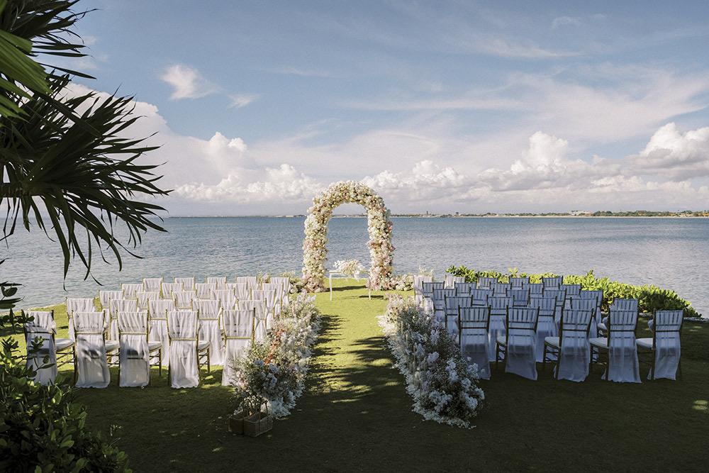 A floral wedding arch and rows of white chairs set on a lawn above the calm water of Jimbaran Bay at Four Seasons Bali