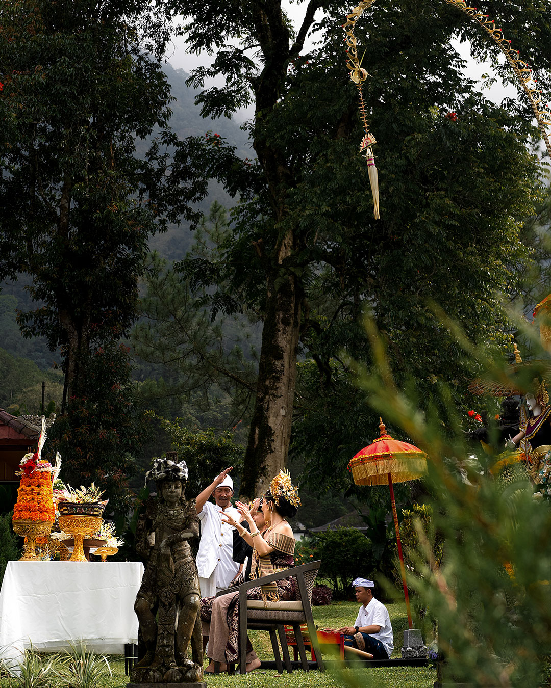 Couple in Balinese ceremonial dress being blessed by a pemangku beside offerings and a red parasol in the gardens at Handara Golf & Resort Bali, Bedugul, Bali