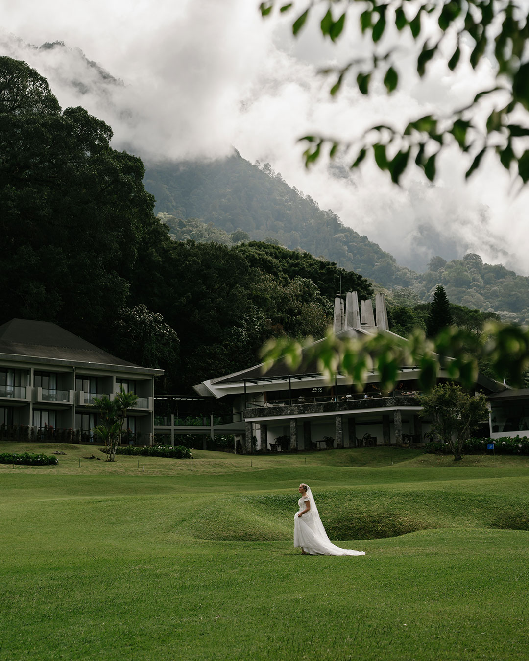 Bride in a long veil standing alone on the fairway below the clubhouse, with cloud on the forested mountain behind, at Handara Golf & Resort Bali, Bedugul, Bali