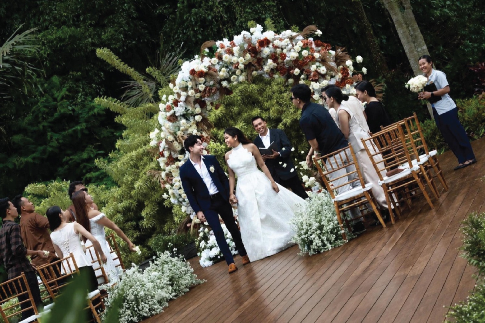 Newlyweds walking back down the timber deck aisle from a white and terracotta floral arch as guests throw petals at HOMM Saranam Baturiti, Bali