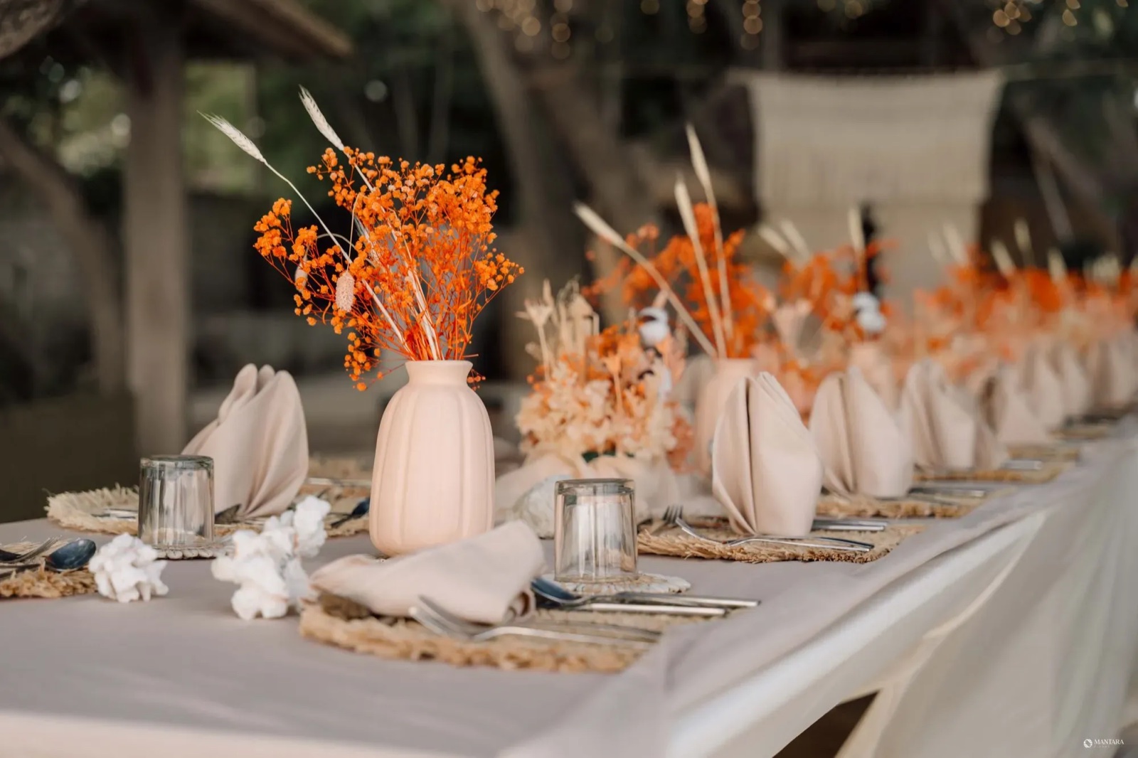 Long wedding table with rattan mats, linen napkins and burnt-orange dried florals under festoon lights at Hotel Lumi Gili Trawangan