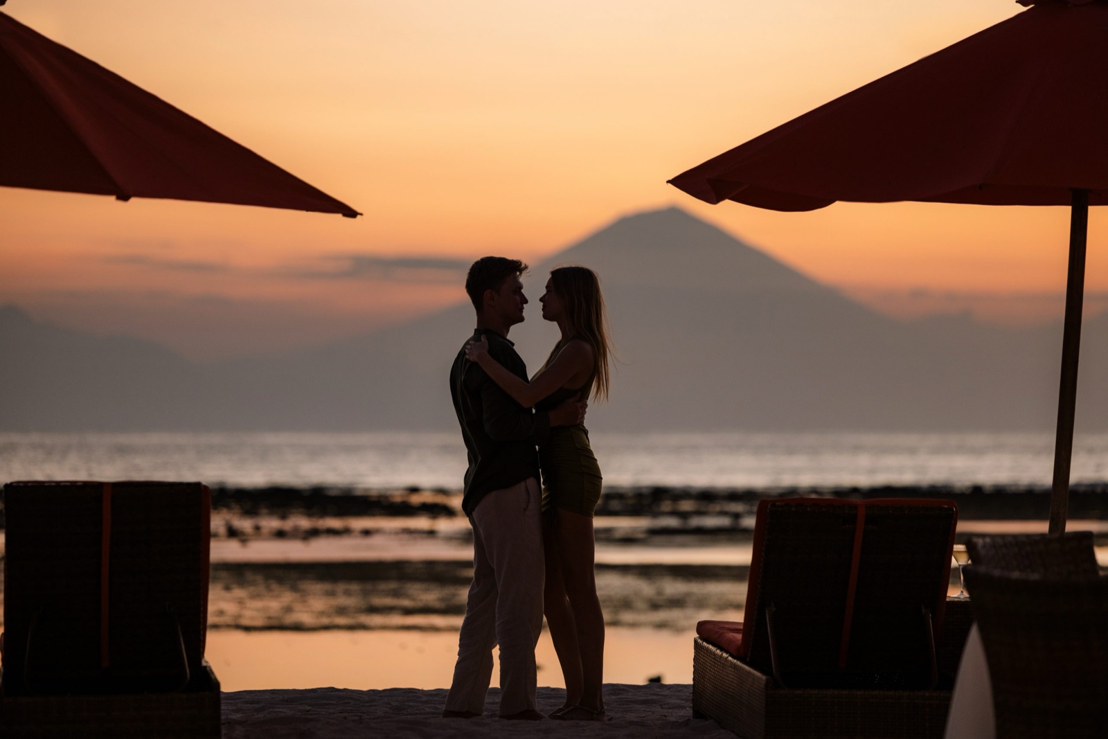 Couple embracing on the sand at sunset with the silhouette of Mount Agung in Bali across the water from Hotel Ombak Sunset, Gili Trawangan