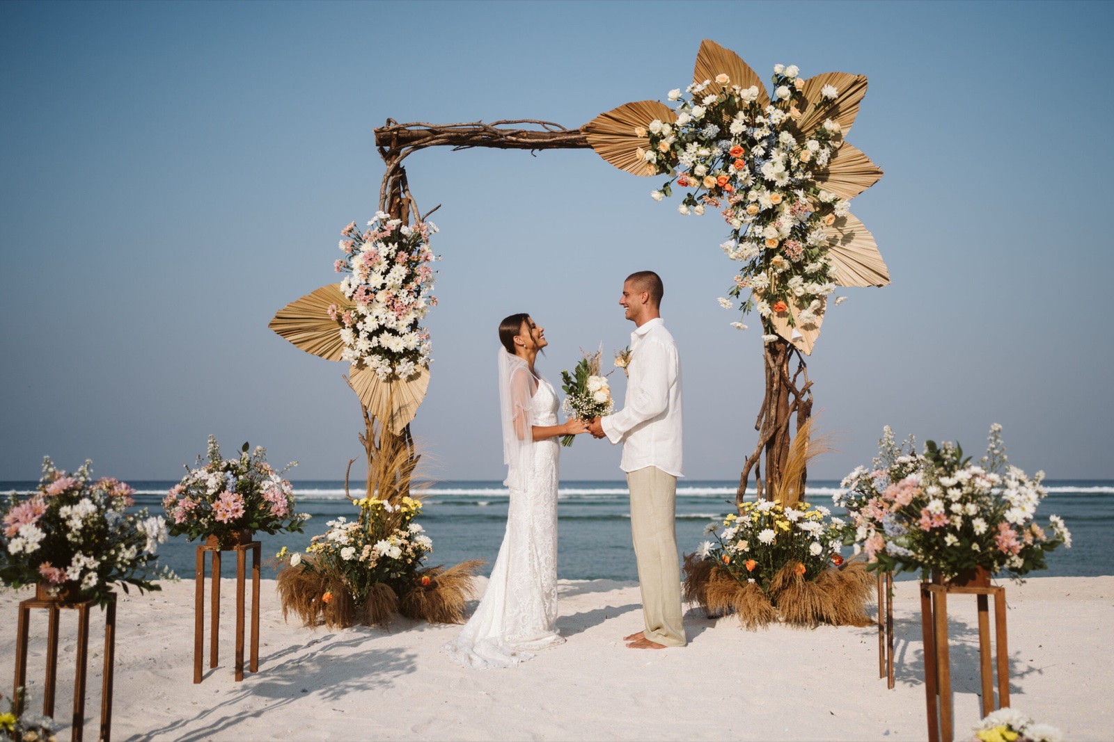 Bride and groom holding hands under a driftwood and dried-palm ceremony arch on the white sand at Hotel Ombak Sunset, Gili Trawangan