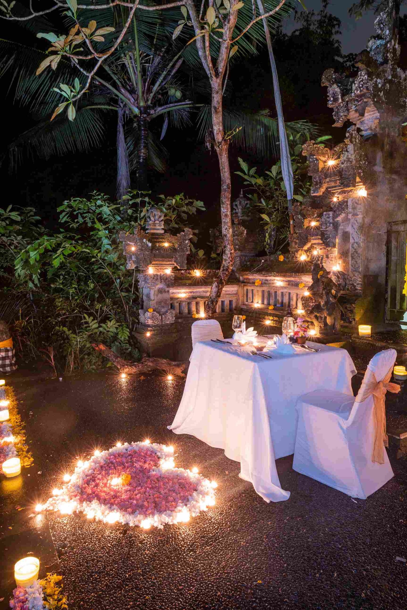 A candlelit dinner for two beside carved temple gates and flower petals in the gardens at night, Hotel Tjampuhan & Spa, Ubud