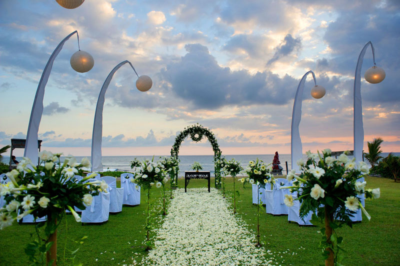 Sunset beachfront lawn ceremony with white florals and a flower-petal aisle facing the ocean at Hotel Tugu Bali, Canggu