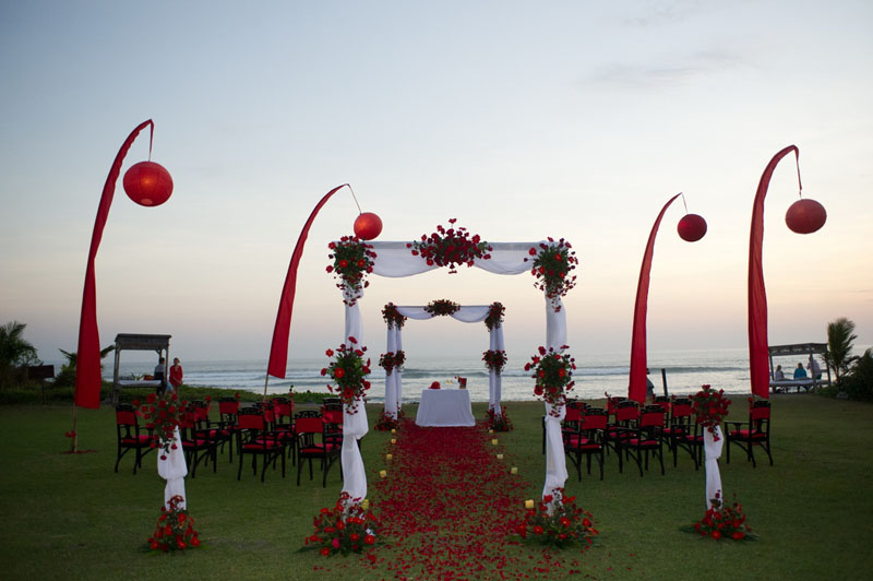 Beachfront wedding ceremony with red-and-white styling and Balinese banners overlooking the sea at Hotel Tugu Bali, Canggu