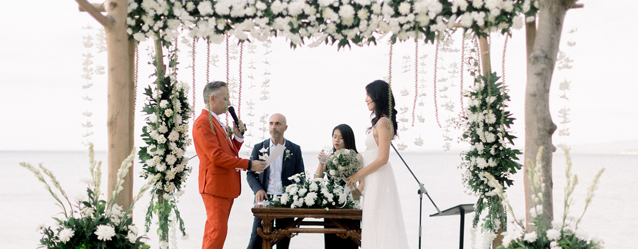 Couple exchanging vows under a timber canopy hung with white blooms and garlands on the sand at Hotel Tugu Lombok, Sire Beach