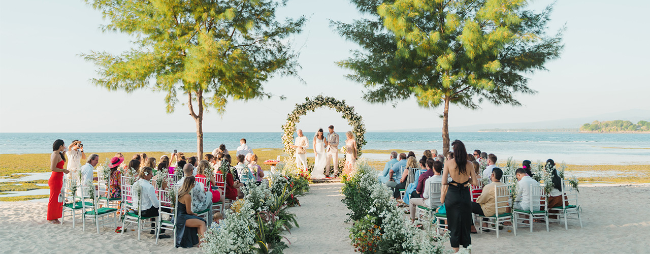 Guests seated on the sand facing a circular floral arch at a beachfront ceremony at Hotel Tugu Lombok on Lombok's northwest coast