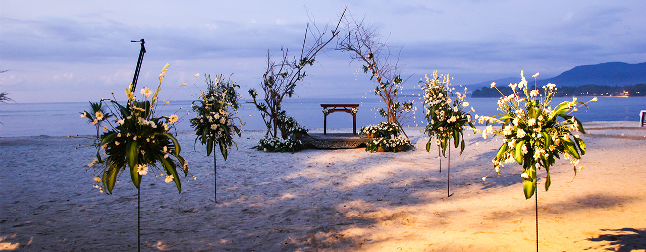 Tall floral installations and a lit ceremony platform set on the sand at dusk at Hotel Tugu Lombok