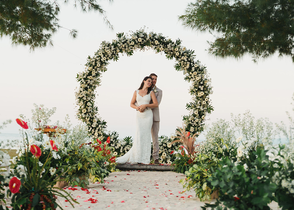 Bride and groom framed by a circular floral arch on the beach at Hotel Tugu Lombok, Tanjung