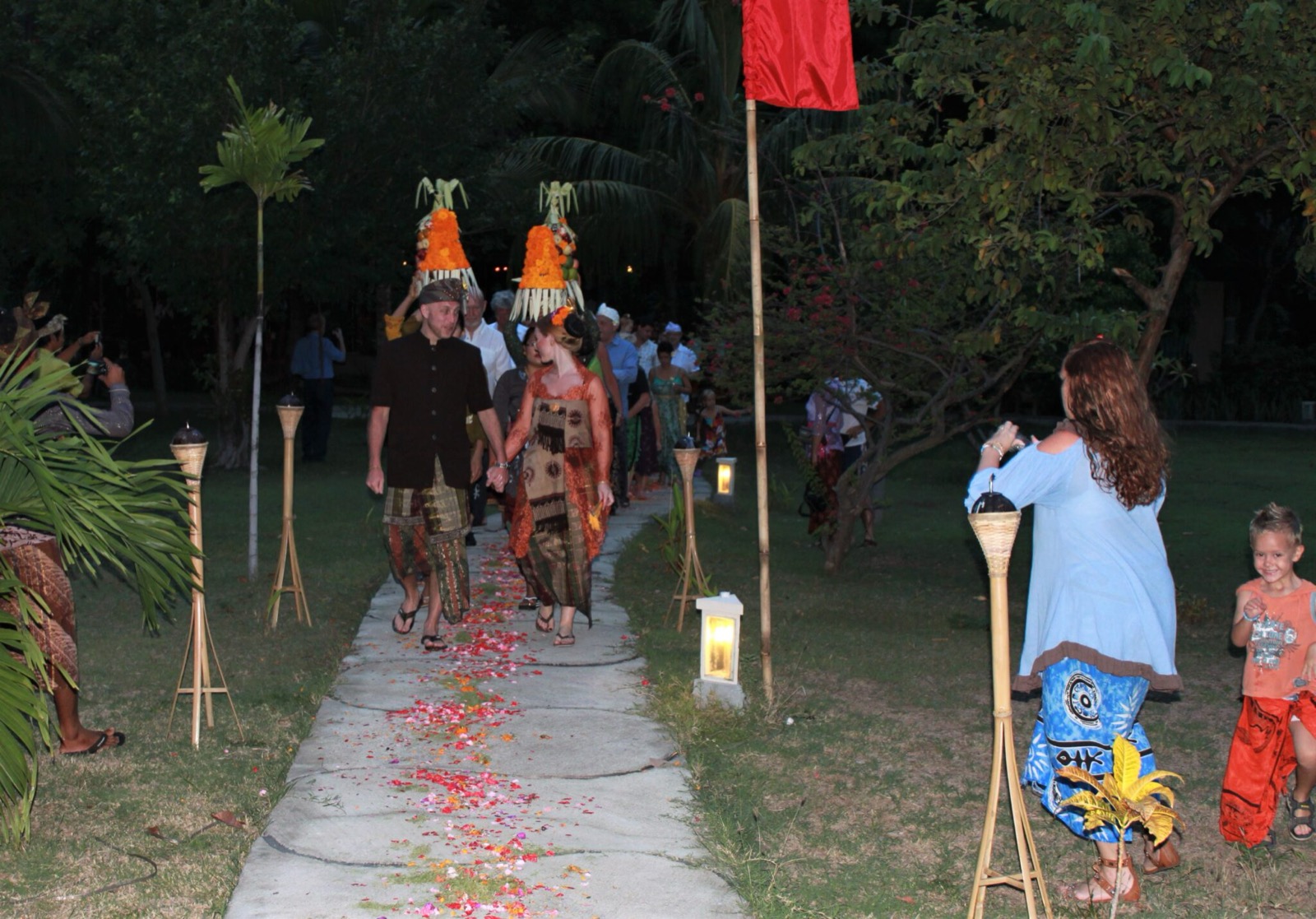 Couple in Balinese dress walking a petal-strewn garden path at night with offerings carried behind them at Hotel Uyah Amed & Spa Resort in Amed, east Bali