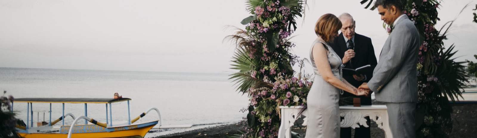 Couple exchanging rings with a celebrant beside a floral arch on the black-sand beach at Hotel Uyah Amed & Spa Resort in Amed, east Bali, with a dive-centre outrigger drawn up behind them