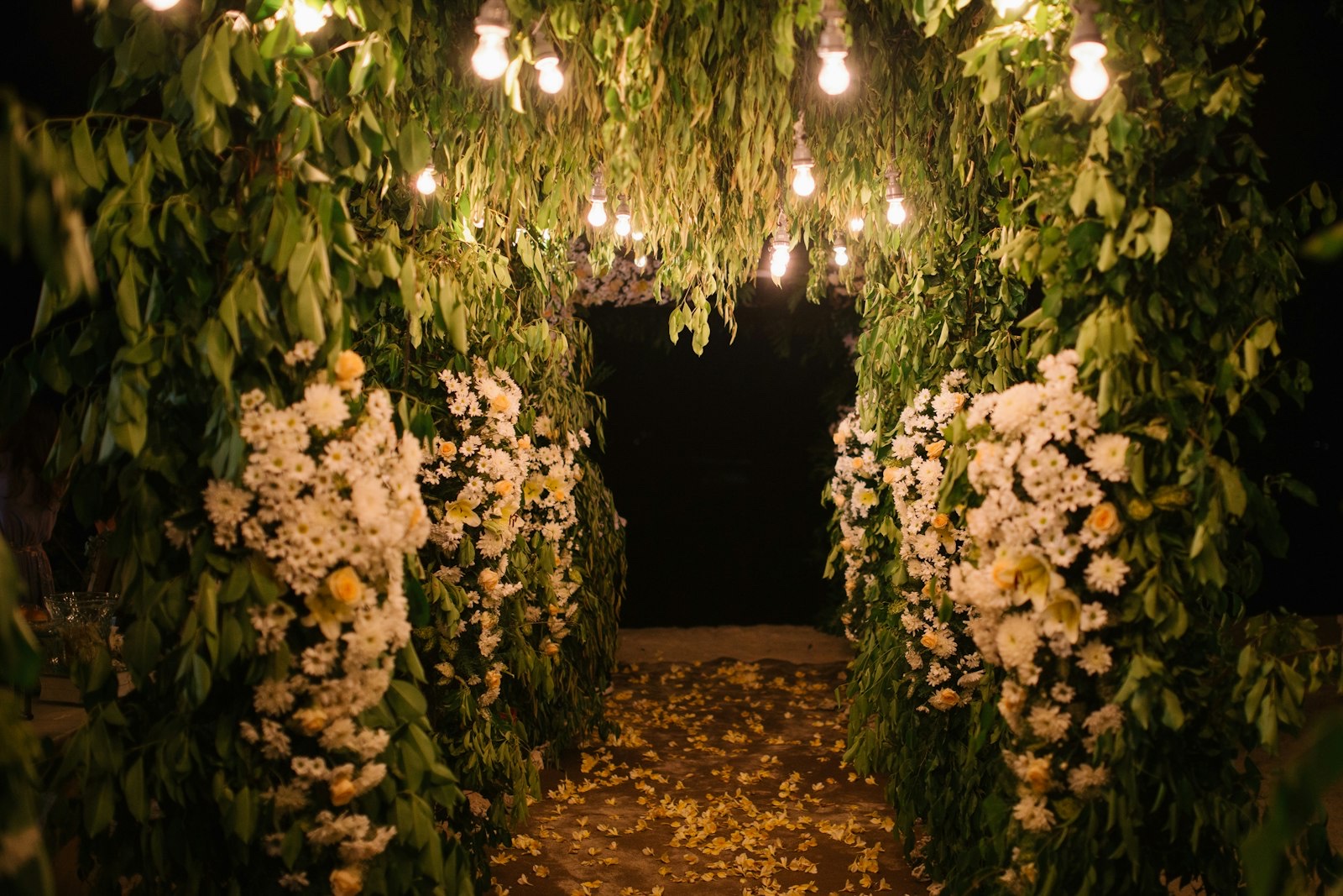 Aisle lined with walls of foliage and white blooms under warm festoon bulbs at an evening wedding at Hotel Vila Ombak, Gili Trawangan