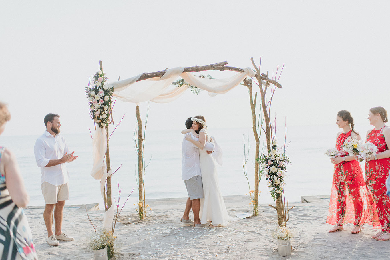 Bride and groom embracing under a driftwood arch draped in cream fabric on Klui Beach, Jeeva Klui Resort, Lombok