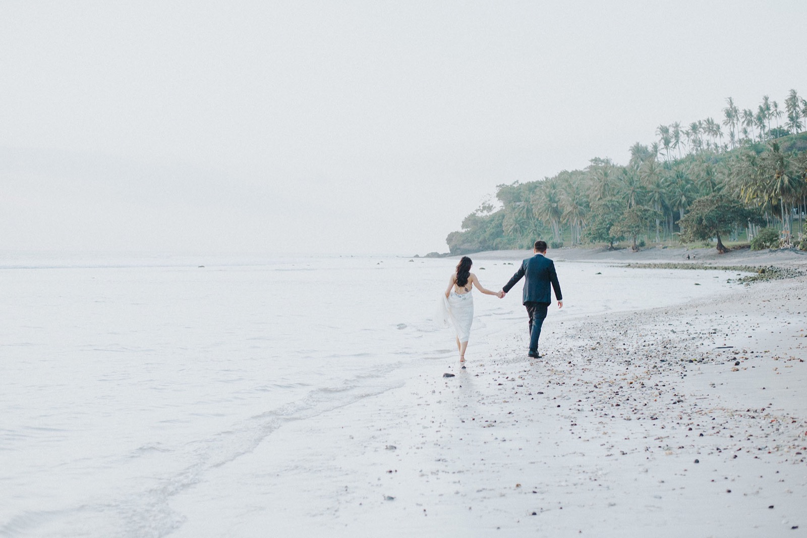 Couple walking the empty shoreline of Klui Beach with the palm-covered headland behind, Jeeva Klui Resort, Lombok