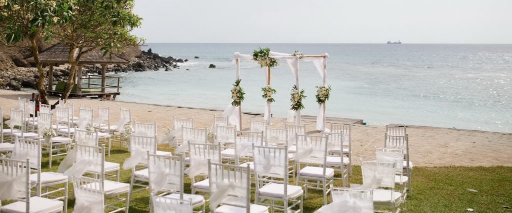 White ceremony chairs on the lawn facing a bamboo arch and the rocky headland at Jeeva Santai Villas, Mangsit, Lombok