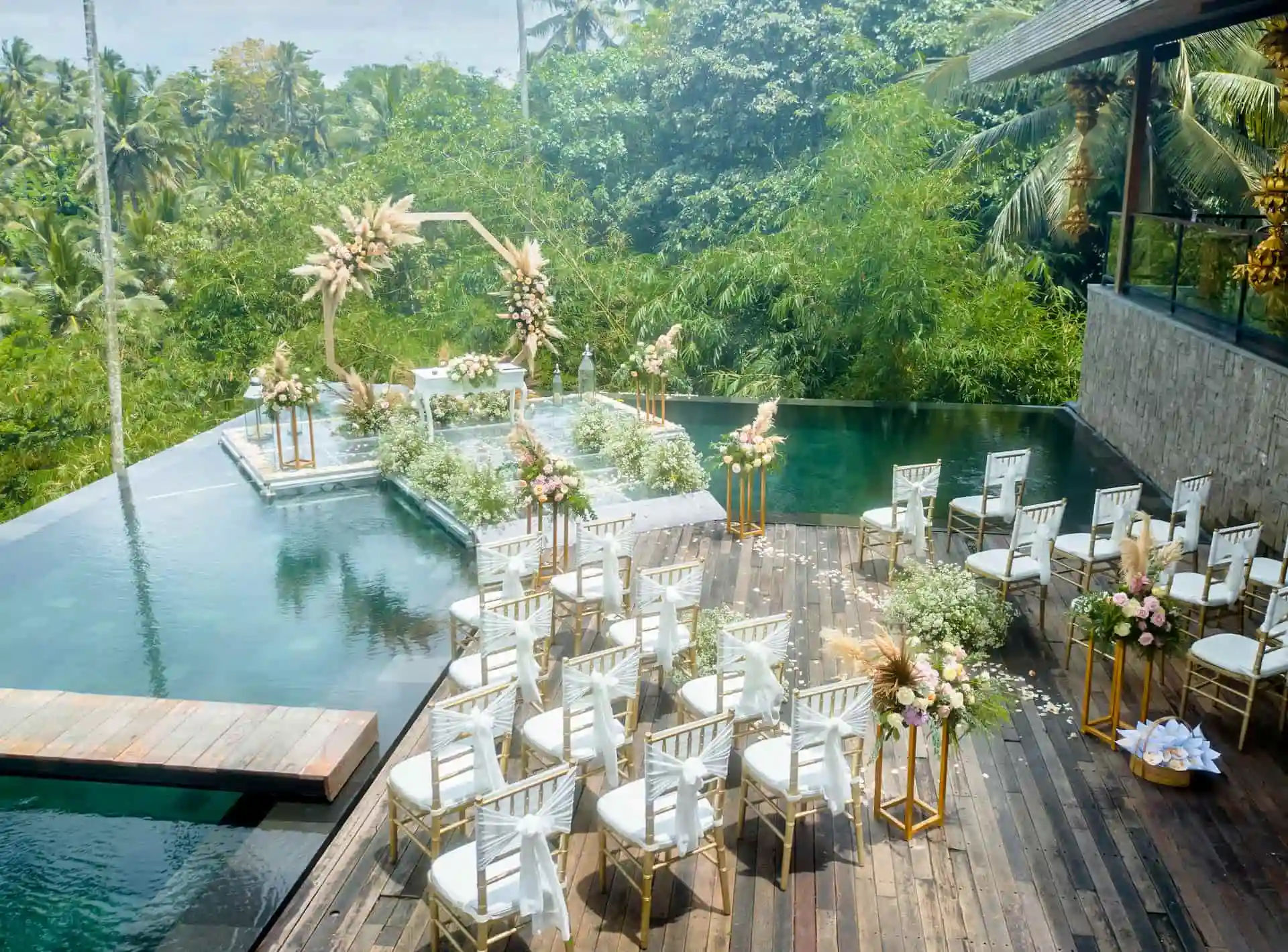 A water wedding set up on the glass stage over the infinity pool at Kaamala Resort Ubud, with gold chairs on the deck and forest beyond