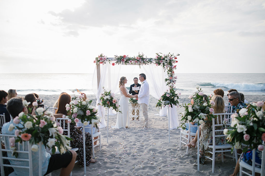 Beach ceremony under a draped floral arch with guests seated on the sand at Katamaran Resort Lombok, Mangsit Beach