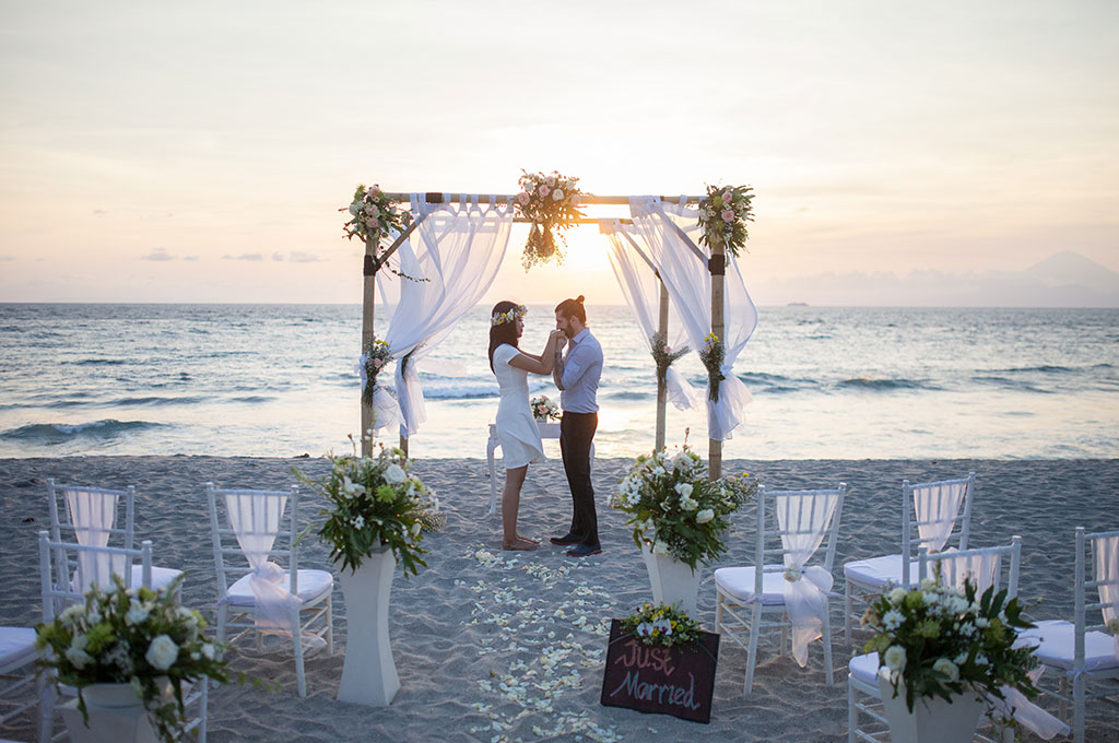 Couple under a bamboo arch on the sand at sunset with Bali's volcano on the horizon, Katamaran Resort Lombok