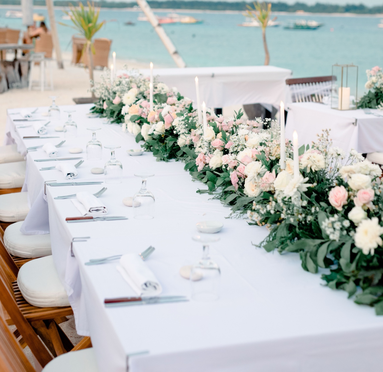 Long wedding table dressed in white with a run of blush and cream florals and taper candles on the sand at Ko-Ko-Mo Gili T Resort, Gili Trawangan