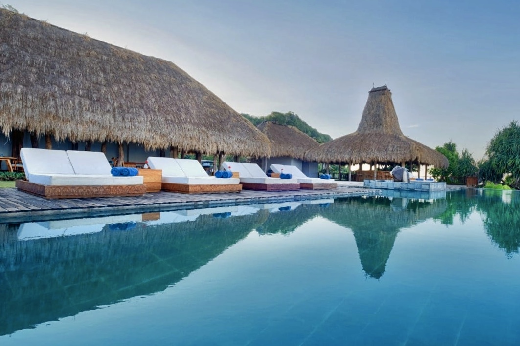 Swimming pool and sun loungers with a tall Sumbanese peaked roof rising behind at Lelewatu Resort Sumba, Sumba