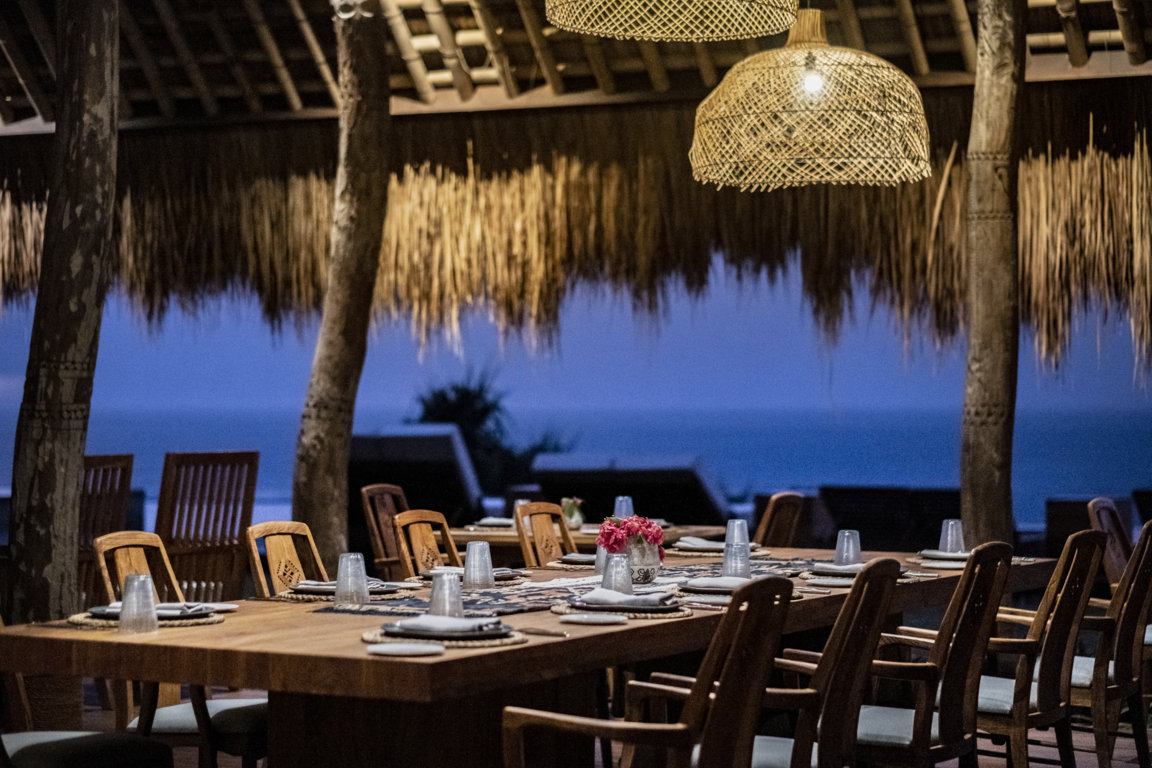 Long dining table set under a thatched roof facing the ocean at dusk at Lelewatu Resort Sumba, Sumba