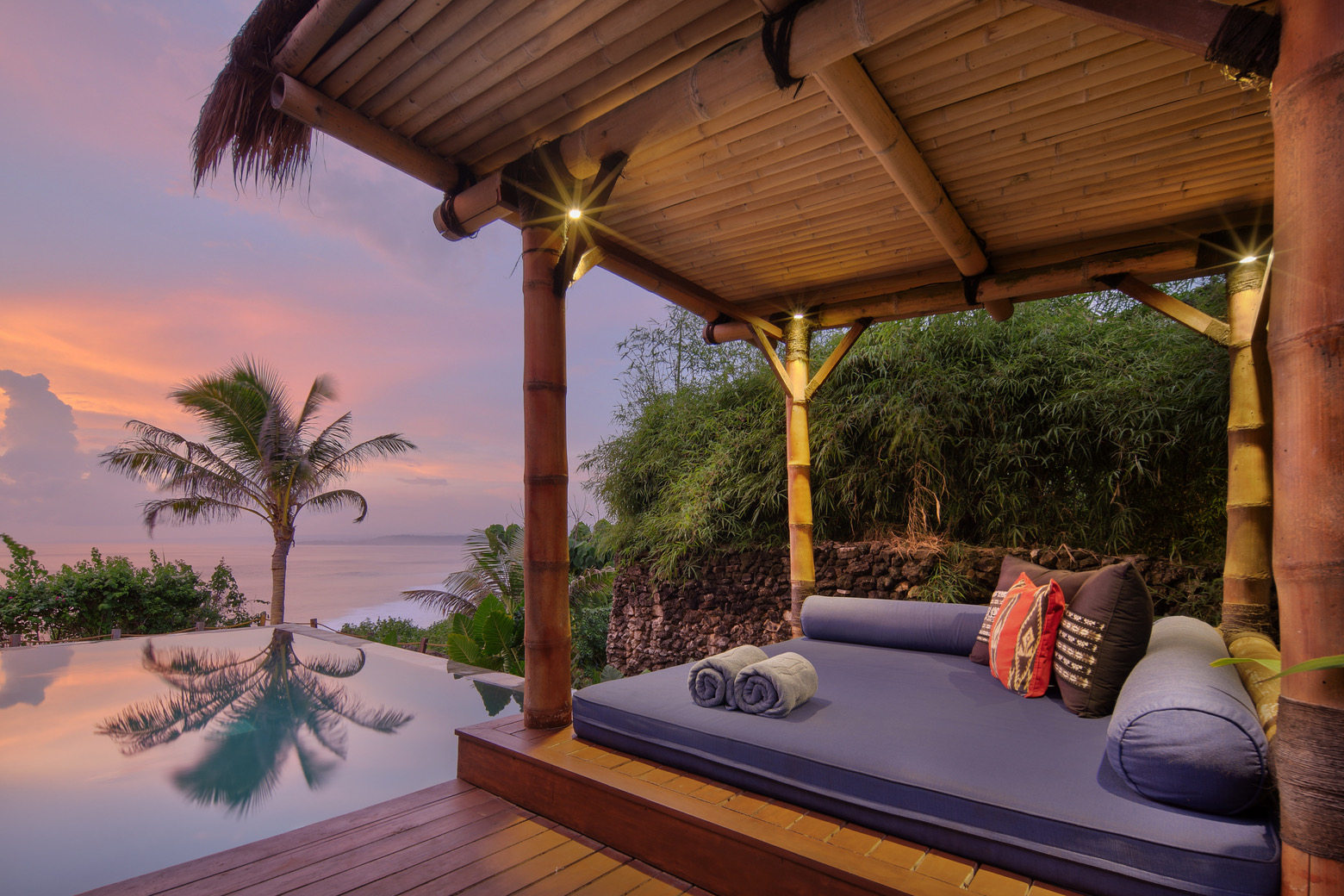 Daybed beside a villa pool at dusk with palms and the sea beyond at Lelewatu Resort Sumba, Sumba