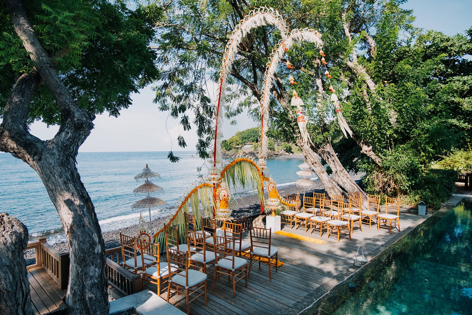 Balinese wedding ceremony set up with marigold arches and Tiffany chairs on the seaside deck above the black pebble beach at Mimpi Resort Tulamben in northeast Bali