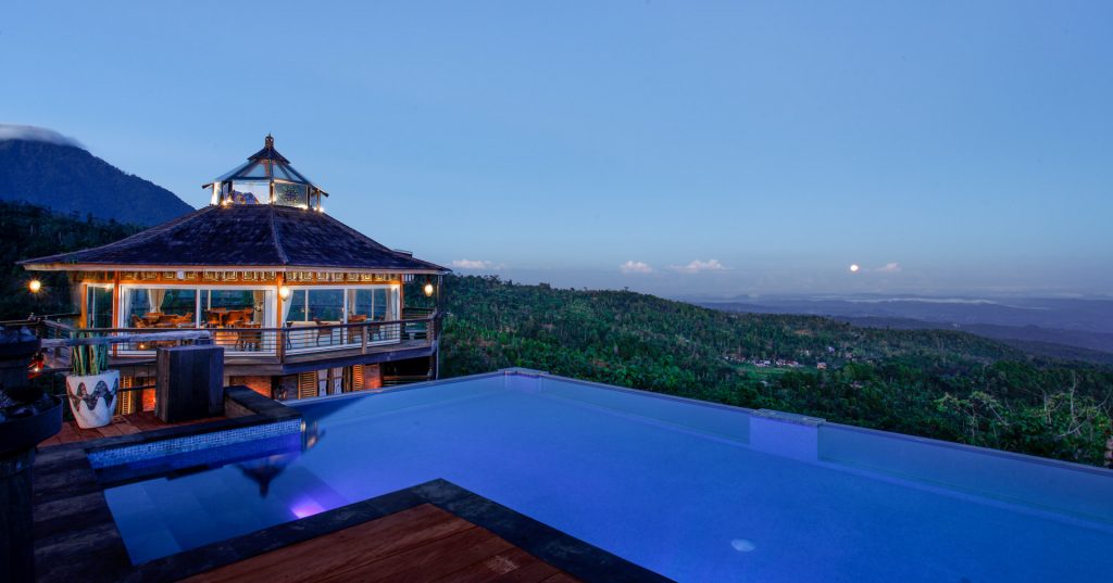 The infinity pool and the lit glass pavilion at blue hour above the forest at Munduk Heaven Luxury Villas, north Bali
