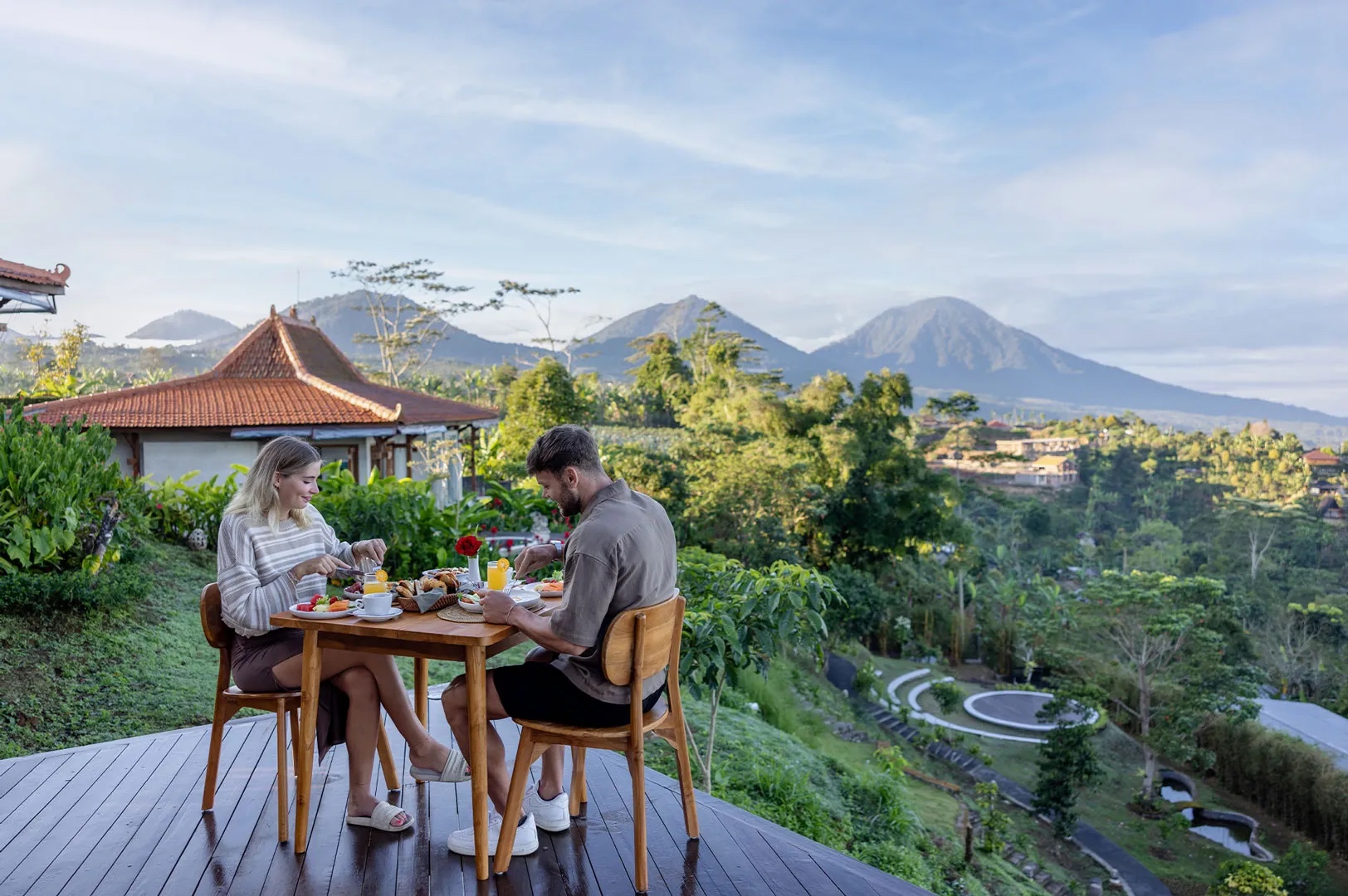 Couple having breakfast on a timber deck with the volcanic peaks behind and the grass amphitheatre below at Munduk Tentrem, Munduk, Bali