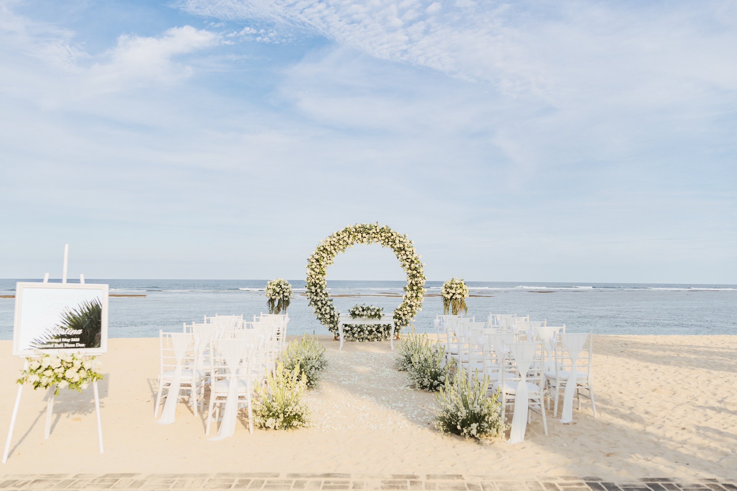 A beach ceremony set with a circular floral arch and white chairs facing the ocean at Novotel Bali Nusa Dua