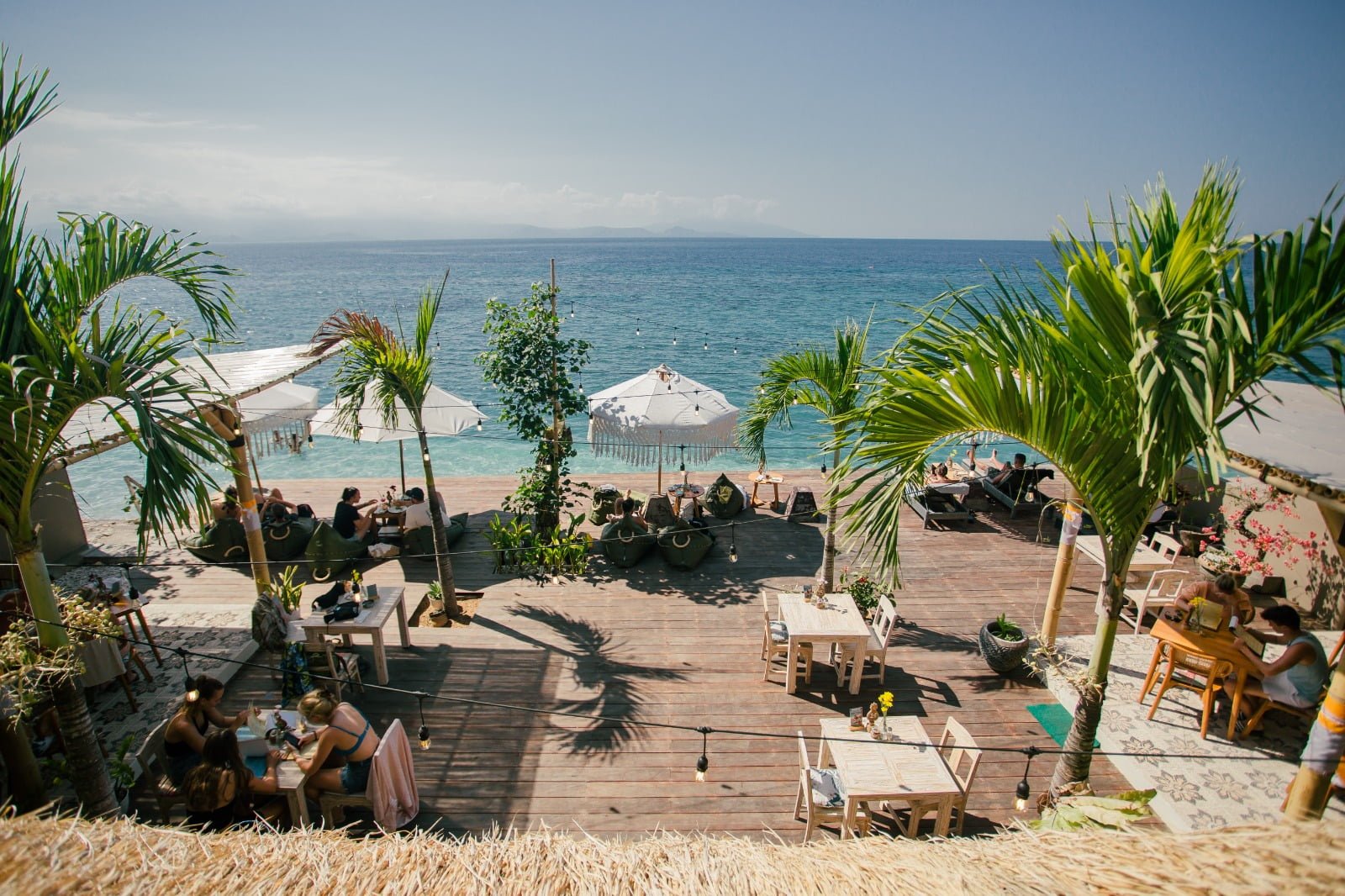 Timber beach deck with bean bags and fringed parasols above the water at Penida Colada, Nusa Penida, Bali