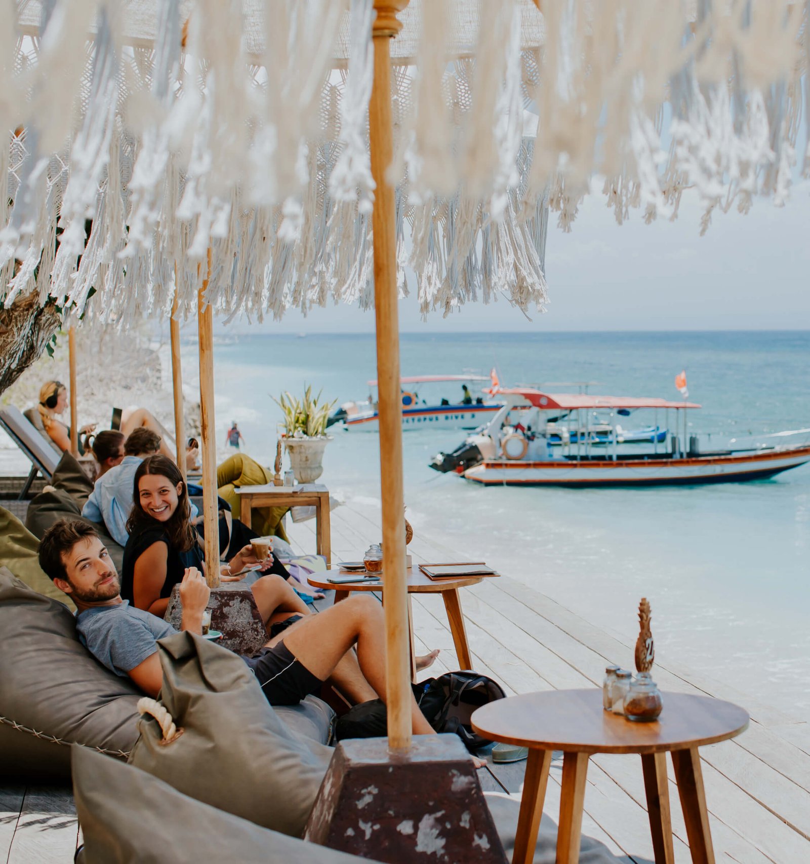 Bean bags under fringed parasols on the deck with jukung boats moored offshore at Penida Colada, Nusa Penida, Bali
