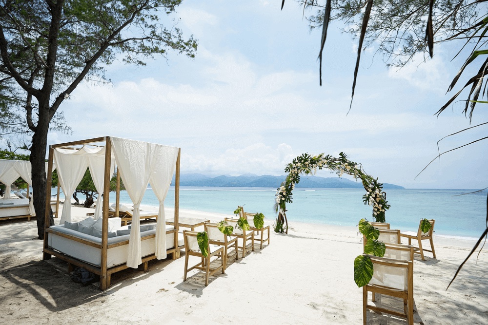 Beach ceremony set with a floral arch, timber chairs and a canopied daybed on the private sand at Pondok Santi Estate, Gili Trawangan