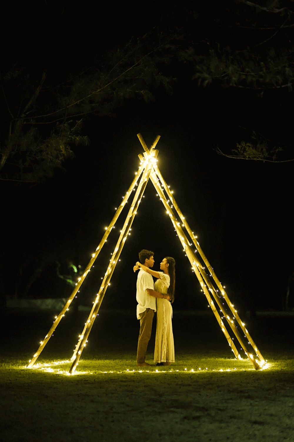 Couple standing under a bamboo A-frame wrapped in fairy lights on the lawn after dark at Pondok Santi Estate, Gili Trawangan