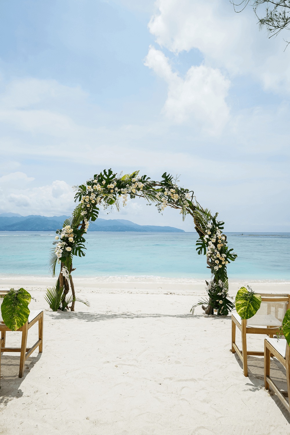 Round floral and monstera arch on the white sand facing the turquoise water and the Lombok hills at Pondok Santi Estate, Gili Trawangan