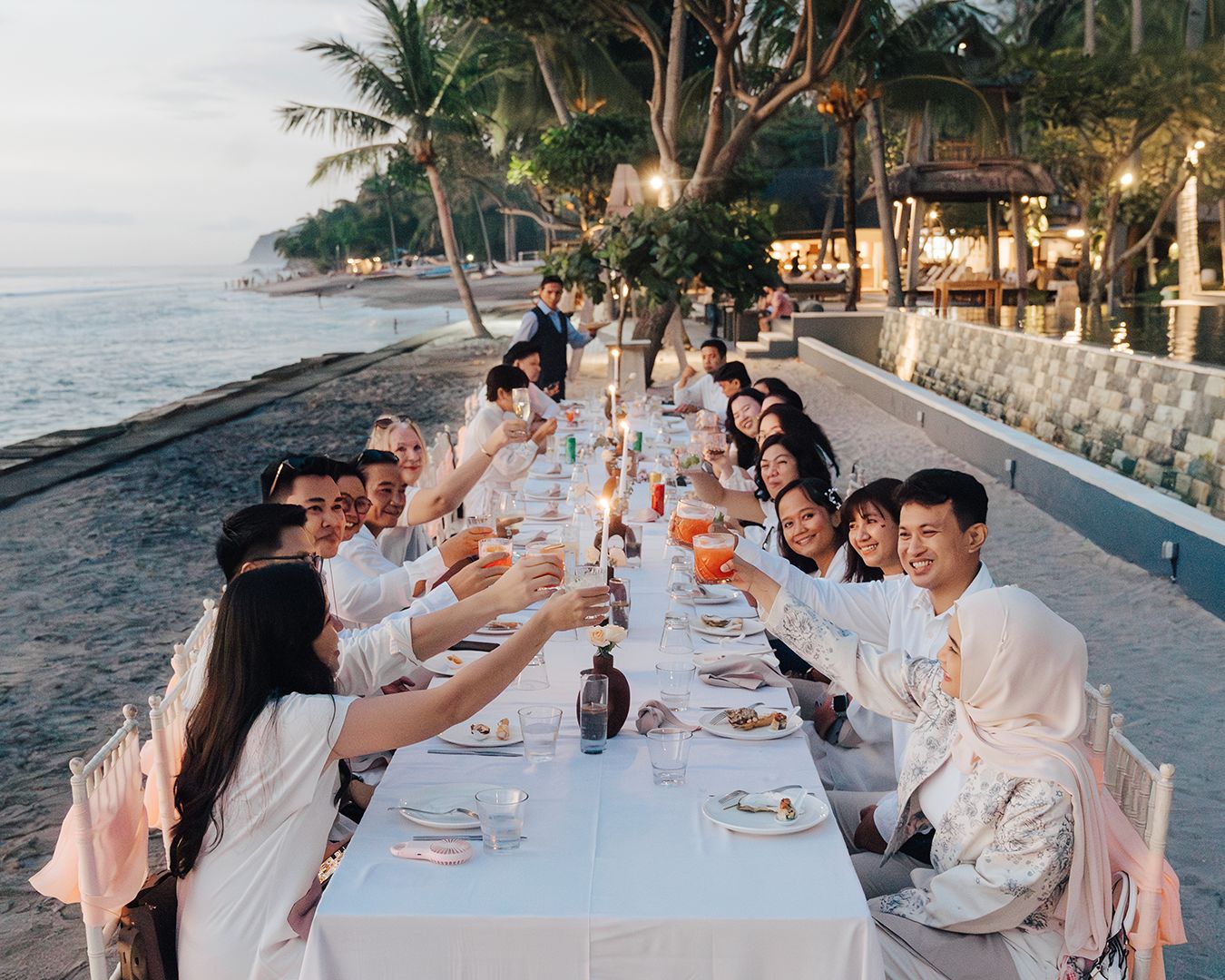 Guests raising a toast along a long white table set on the sand at dusk at Qunci Villas on Lombok's west coast