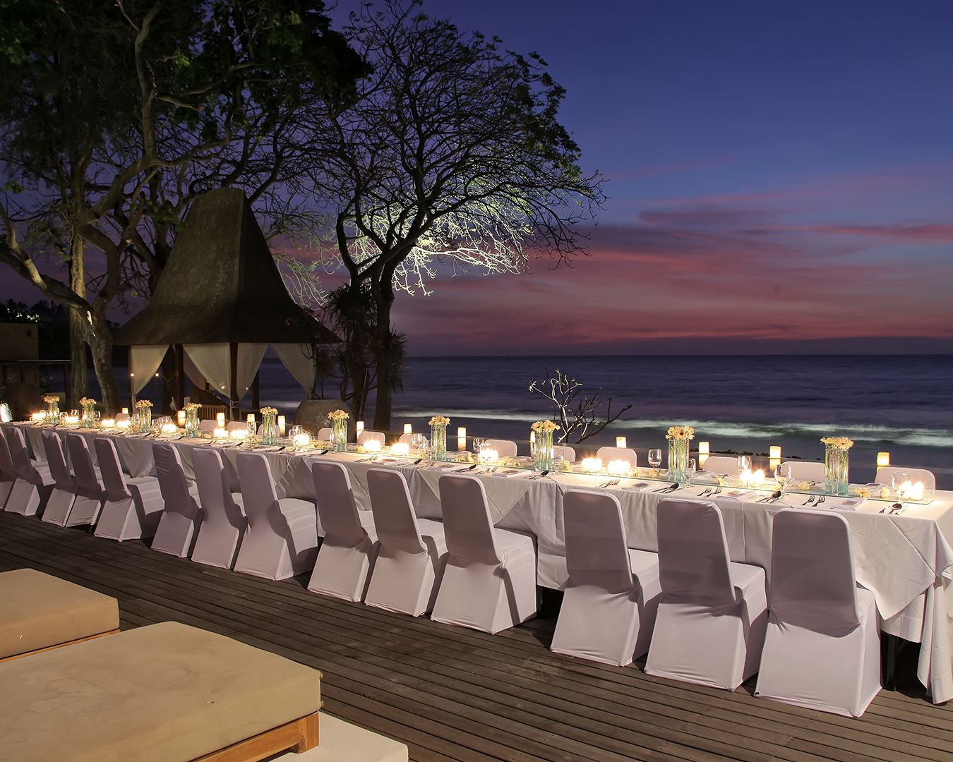 Candlelit long table laid on the beachfront deck at twilight beside a thatched berugak at Qunci Villas, Lombok