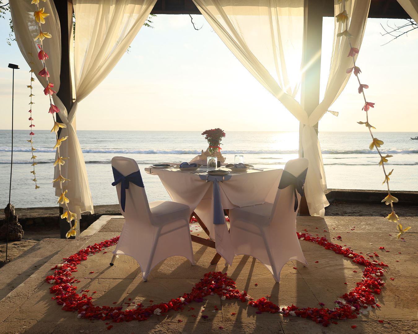 Table for two set under a draped berugak facing the sea with a heart of red petals on the terrace at Qunci Villas, Lombok