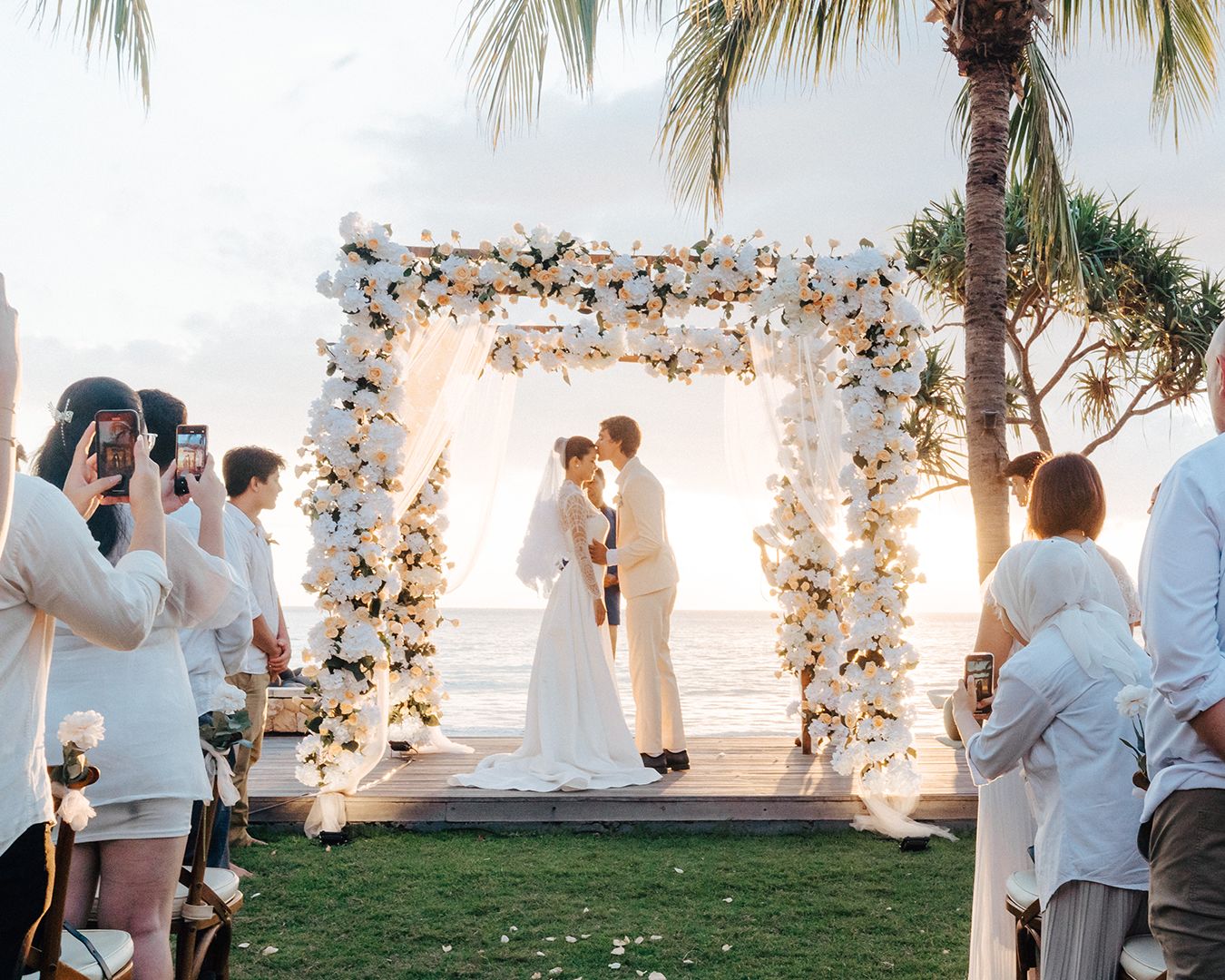 Couple kissing under a white and peach floral arch on the beach deck at sunset while guests watch, Qunci Villas, Lombok