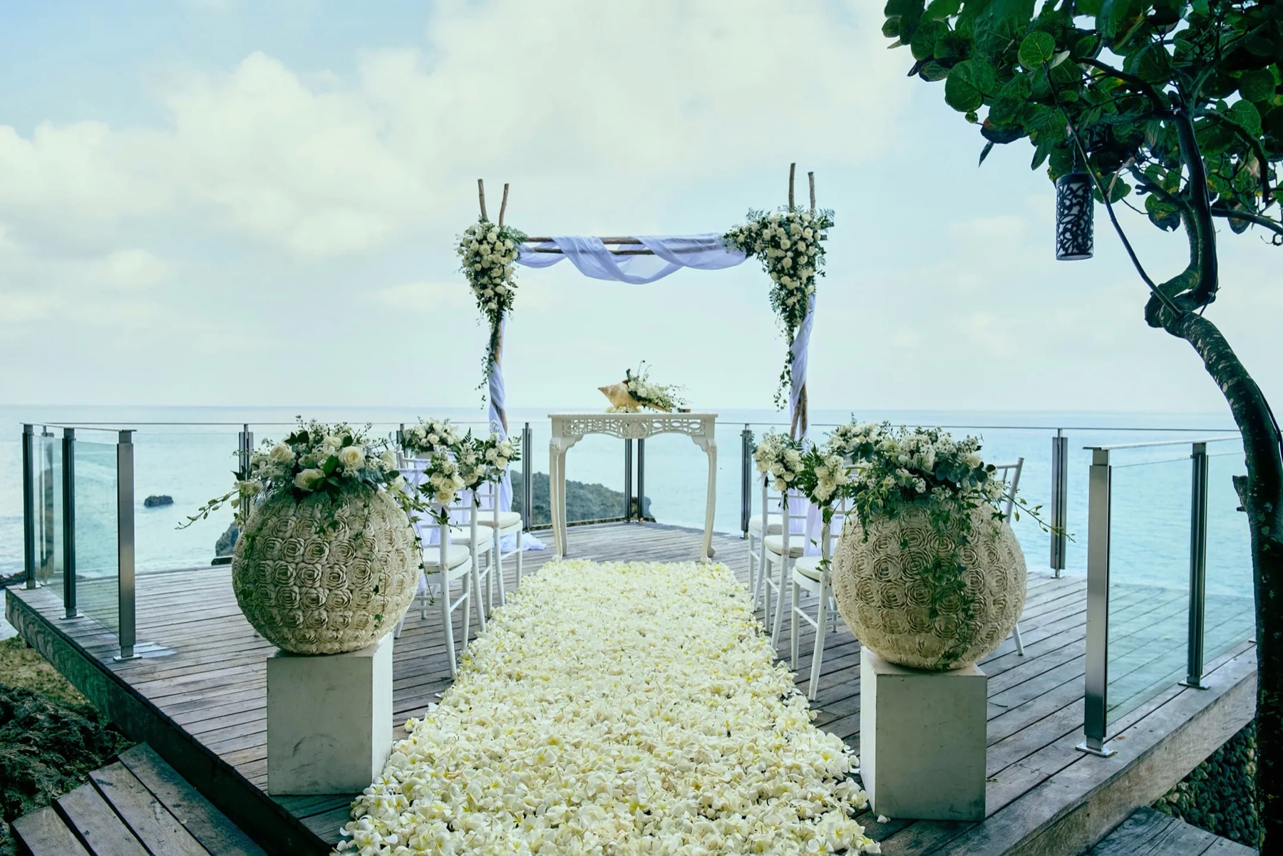 A wedding aisle of white petals on a timber deck at Rock Bar, AYANA, framed by a flower arch and glass railings above the Indian Ocean